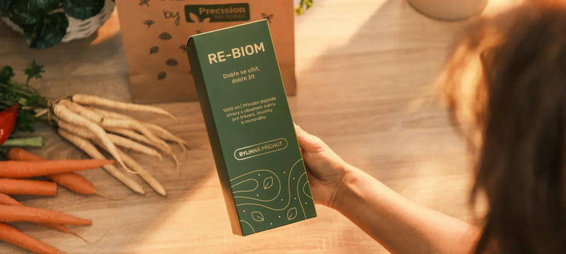 Ženská ruka drží zelenou krabičku s postbiotiky Re-Biom na světlém pozadí.